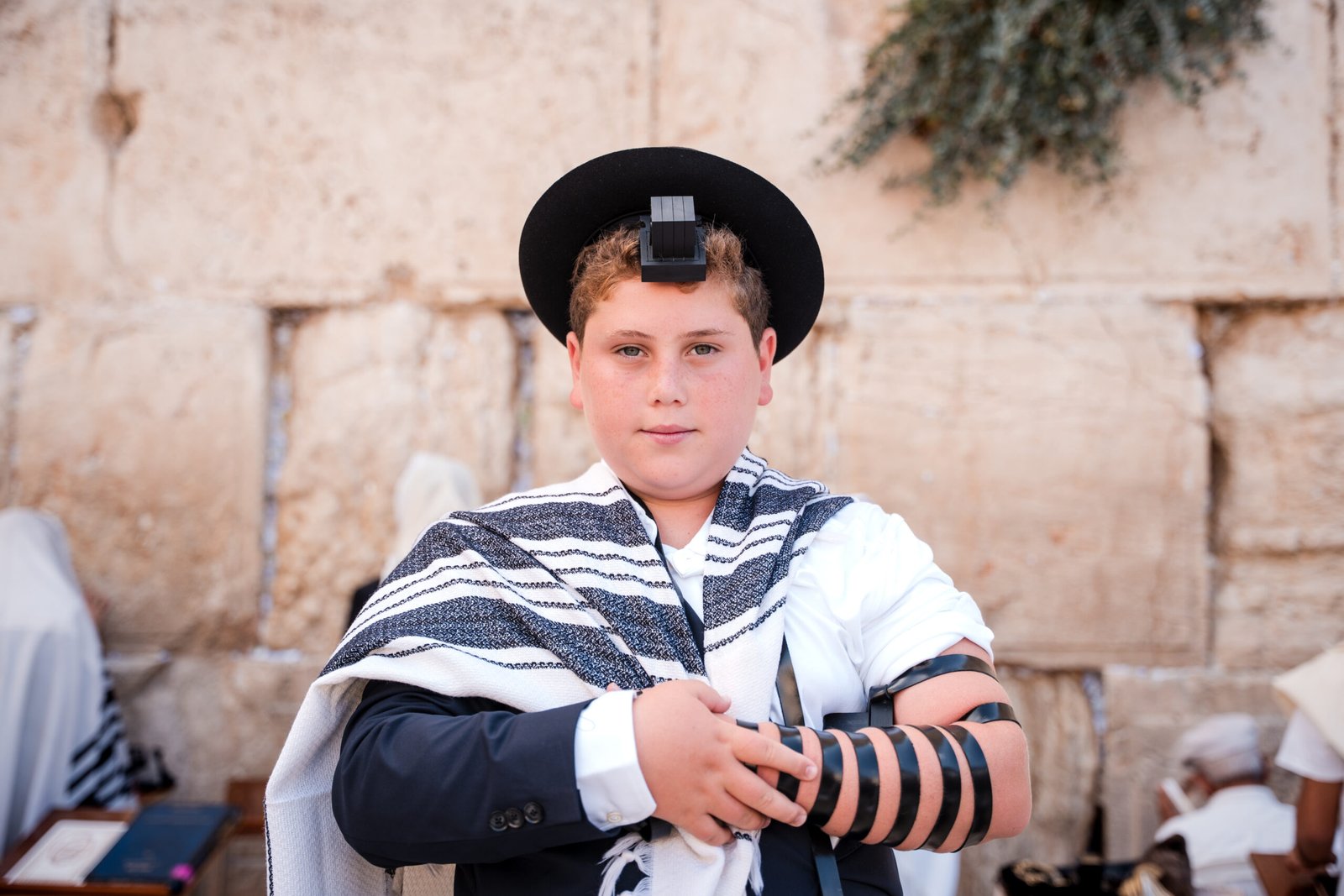 A solemn young Jewish boy, possibly celebrating his Bar Mitzvah, looking directly at the camera. He is wearing a black hat, a tallit (prayer shawl) draped over a suit jacket,