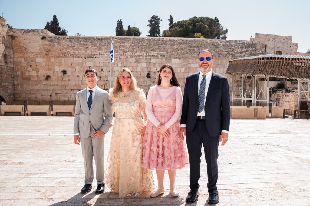 Family of four standing in front of the Western Wall, including a boy in a grey suit, a woman in a light floral gown, a girl in a pink dress, and a man in a dark suit, all smiling in bright sunlight