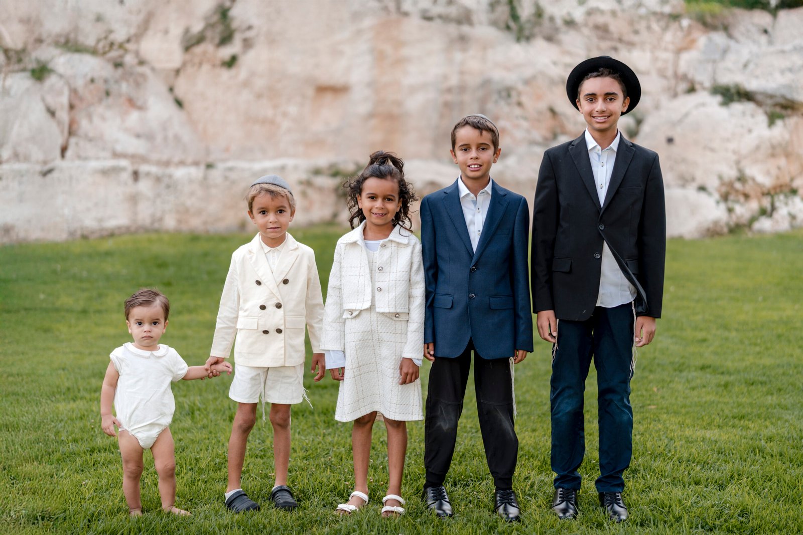 A group of children dressed in formal attire — boys in suits and a girl in a white dress — standing on grass with an old stone wall in the background.