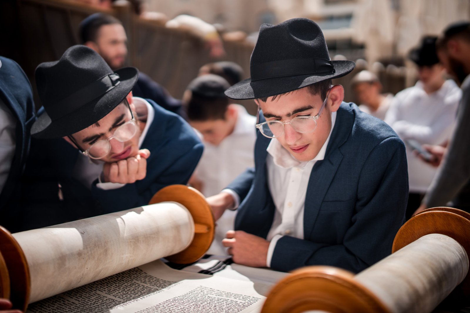 Two young Jewish boys or Bar Mitzvah celebrants, wearing dark suits, white shirts, glasses, and black fedora hats, intently reading from an open Torah scroll.