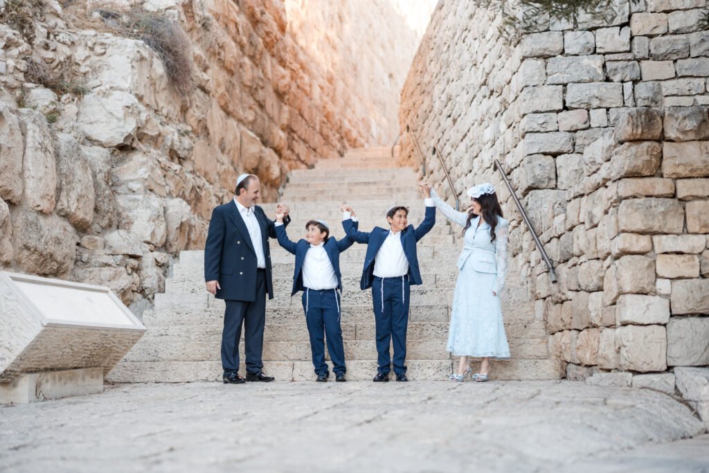 A Jewish family, likely celebrating a Bar Mitzvah, poses on ancient stone steps outdoors between two massive retaining walls in Jerusalem. The father, wearing a dark suit, and the mother, in a light blue dress and white head covering