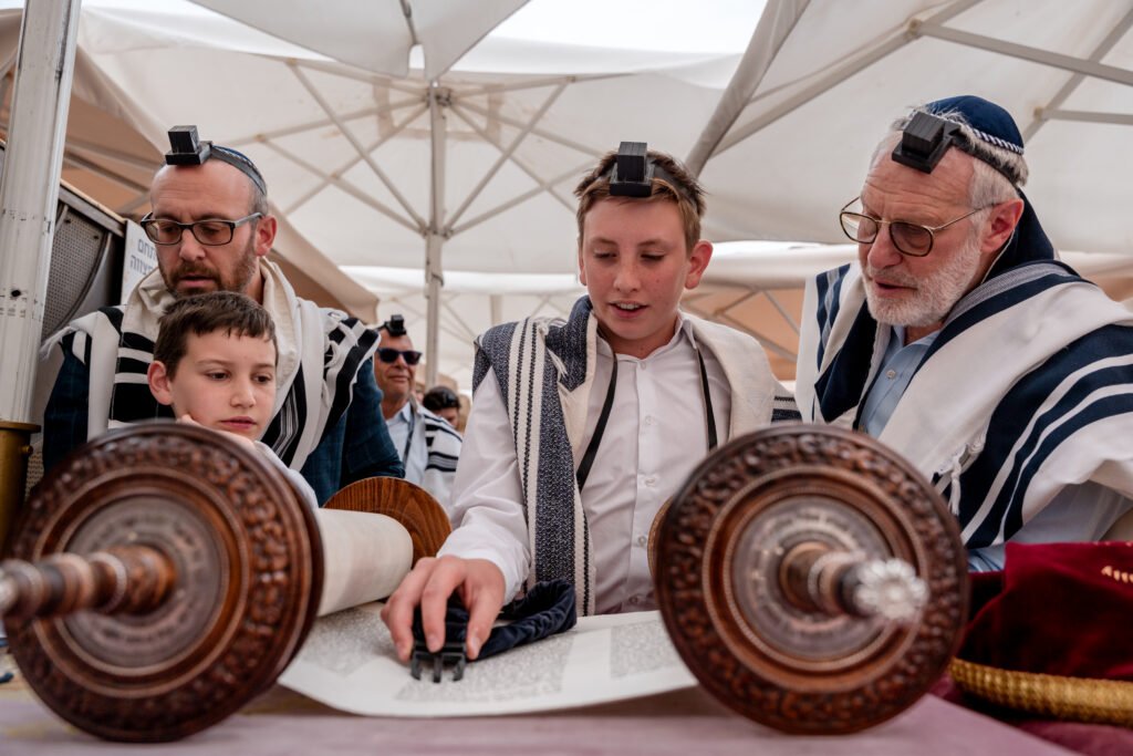 A young Bar Mitzvah boy, wearing tefillin and a tallit (prayer shawl), reads from an open Torah scroll during a service outdoors, possibly at the Western Wall. He is flanked by two older men, likely his father and grandfather, who are also wearing tefillin and tallitot, attentively watching him read.