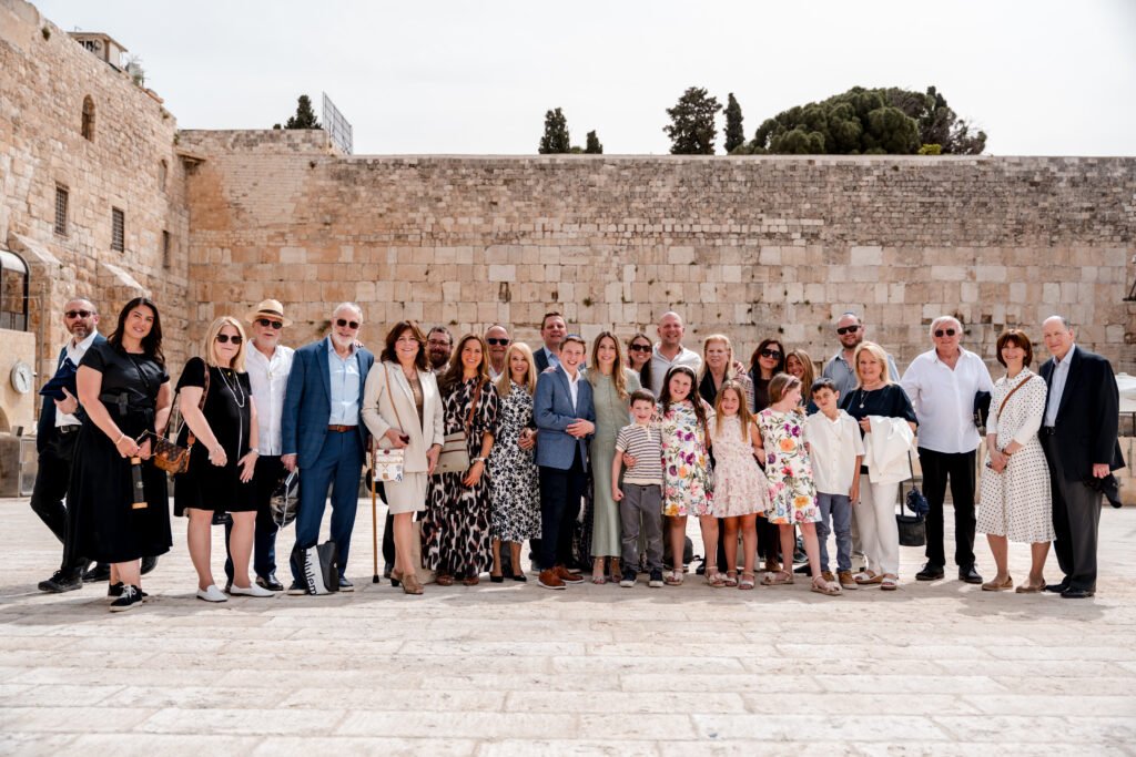Large family group standing together in front of the Western Wall, including adults and children dressed in formal and semi-formal outfits, smiling for a group photo outdoors